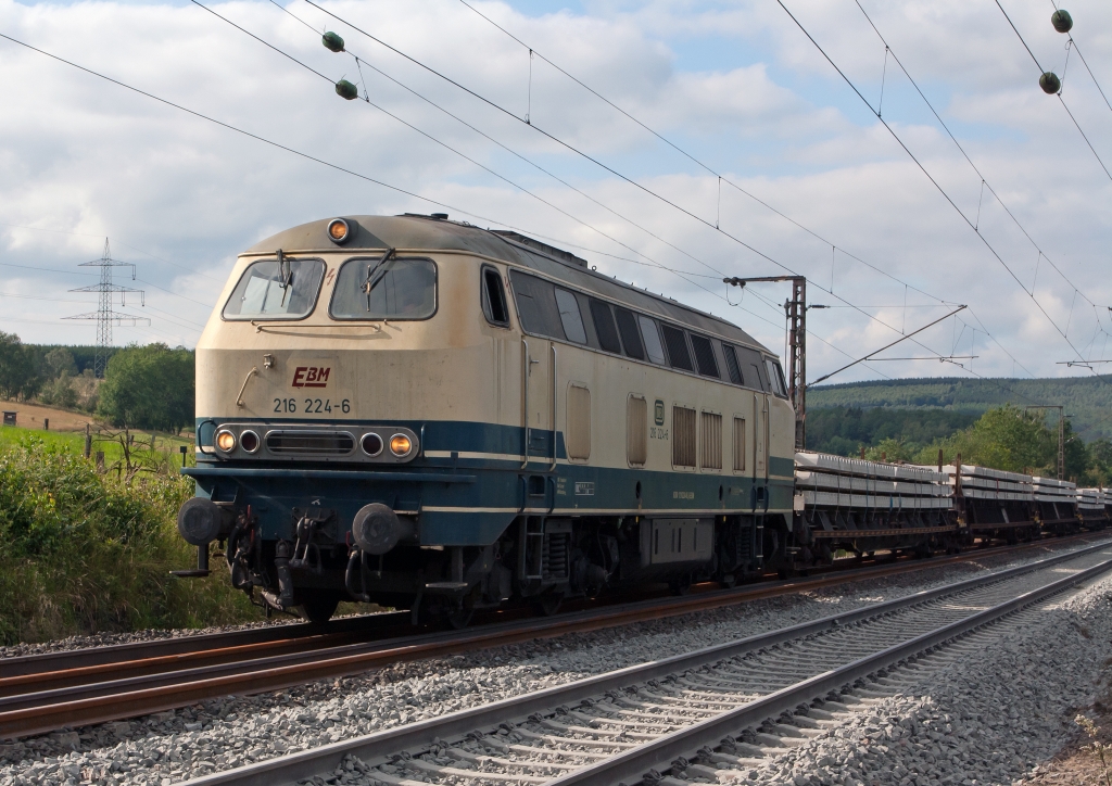 Die 216 224-6 der EBM Cargo (Gummersbach) kommt mit einem Schwellenzug am 29.07.2011 von Siegen �ber das Baugleis in Wilnsdorf-Rudersdorf (KBS 445). Die Lok wurde 1968 bei Krupp unter der Fabriknummer 4885 gebaut.
Die Baureihe V 160 (ab 1968: Baureihe 216) war die erste Variante der V 160-Familie der Deutschen Bundesbahn, die im Nachkriegs-Neubauprogramm als einmotorige Gro�diesellokomotive f�r den mittelschweren Streckendienst projektiert wurde.
Angetrieben wird die Lok vom Dieselmotor MTU 16 V 538 / 653 TB 10 mit 16 Zylindern und einer Leistung von 1900 PS bei 1500/min. Die Leistung gelangt �ber ein hydraulisches Getriebe mit zwei Wandlern und einer Kupplung und Gelenkwellen zu den Radsatzgetrieben in den beiden zweiachsigen Drehgestellen. Zus�tzlich zum Fahrdieselmotor ist ein kleiner Hilfsdieselmotor vorhanden, welcher beim Aufr�sten -- insbesondere zum Betrieb des Dampfheizkessels -- und als Kompressor zur Luftversorgung genutzt werden kann.