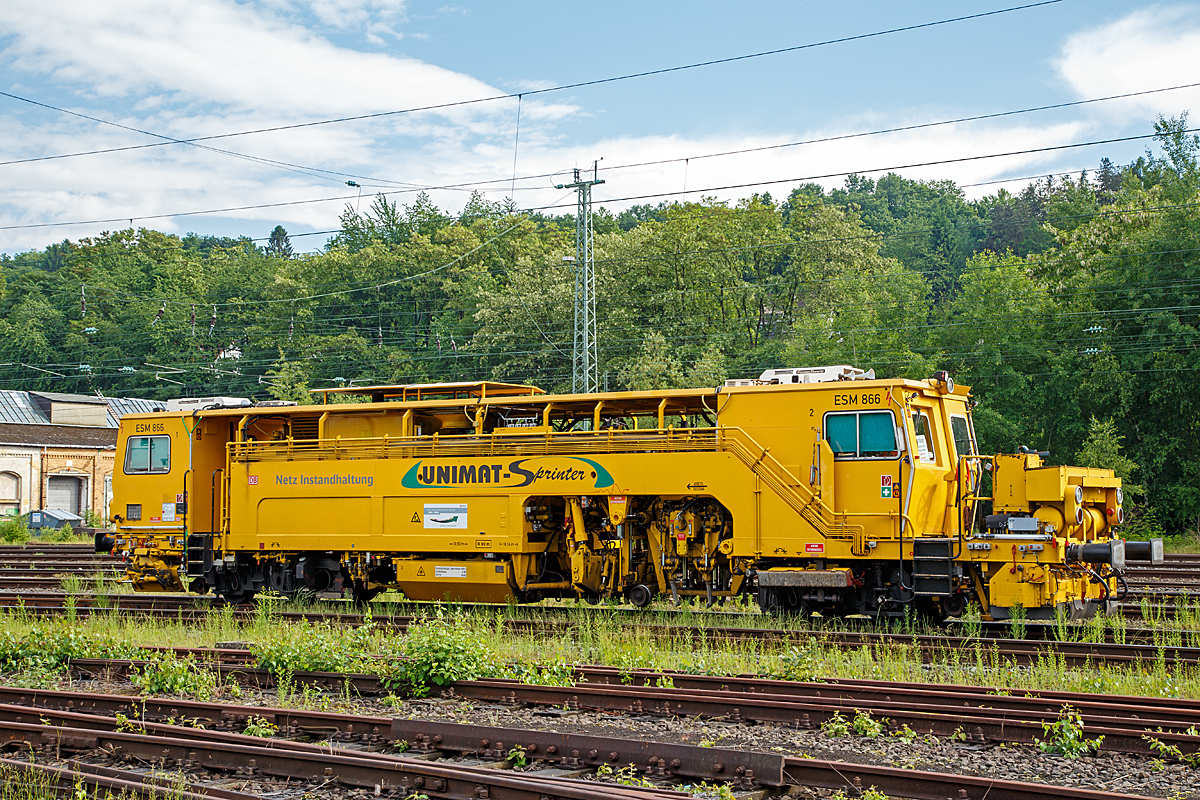 
Die Universal-Stopfmaschine Unimat-Sprinter ESM 866 der DB Netz AG (Schweres Nebenfahrzeug Nr. 97 43 52 006 17-1) steht am 04.06.2016 im Betzdorf/Sieg im Abstellbereich. Der Unimat-Sprinter ist eine von Plasser und Theurer entwickelte Stopfmaschine für Gleise, Weichen und Kreuzungen, speziell konzipiert für die rasche und präzise Behebung von Einzelfehlern. Daher auch die Abkürzung ESM = EinzelfehlerStopfMaschine.

Die Stopfmaschine (ESM – 08 – 275 / Sprinter) wurde 2000 von Plasser & Theurer unter der Fabriknummer 1169 gebaut und an die DB Netz AG geliefert.

Die typische Länge eines Einzelfehlers beträgt 9 bis 15 Schwellen, er kann sich in Einzelfällen aber bis zu 100 m weit erstrecken. Die Ursachen liegen nicht im direkten  Zusammenhang  mit  Nivellier-  und  Richtarbeiten oder Zugbelastungen, sondern vielmehr in punktuellen, scharf   abgegrenzten Schwächen im Unterbau, Frostschäden usw.
Da dieser Einzelfehler in einem völlig intakten Abschnitt liegt,  der keiner durchgehenden Behandlung bedarf, ist es viel zu aufwendig, solche Abschnitte zur Gänze zu bearbeiten. Die Behebung eines Einzelfehlers ist aber unumgänglich, da nicht  nur Gründe wie kostspielige Langsamfahrstellen dafür sprechen, sondern auch die Sicherheit des Fahrweges garantiert werden muss.

Der UNIMAT SPRINTER ist eine speziell für die Einzelfehlerbehebung entwickelte Stopfmaschine. Sie ist mit zwei Weichenstopfaggregaten mit jeweils vier Schwenkpickeln zur universellen Bearbeitung von Strecken und Weichen  ausgestattet. Das kombinierte Hebe- und Richtaggregat besteht aus einem Hebehaken, einer Richtrolle und einer Rollhebezange pro Schienenstrang. Für eine schnelle Überstellung zur Baustelle ist die Maschine mit einem hydrodynamischen Antrieb für eine Höchstgeschwindigkeit von 100 km/h ausgelegt. Flankenpflüge, die im Bereich des vorderen Drehgestells montiert sind und eine Kehrbürste am hinteren Ende der Maschine, sorgen nach den Stopfarbeiten für  die Herstellung des geforderten Schotterprofils.

Technische Daten der UNIMAT SPRINTER:
Spurweite: 1.435 mm
Achsanzahl: 4 (in 2 Drehgestellen)
Achsabstand im Drehgestell: 1.520 mm
Drehzapfenabstand: 10.500 mm
Länge über Puffer: 18.140 mm
Eigengewicht: 54t
Anhängelast: 40 t
Höchstgeschwindigkeit: 100 km/h
Zugelassen zur Mitfahrt: 4 Pers.
Kleinster befahrbarer Kurvenradius:  R 90 m      
Zugelassen für Streckenklasse: A und höher