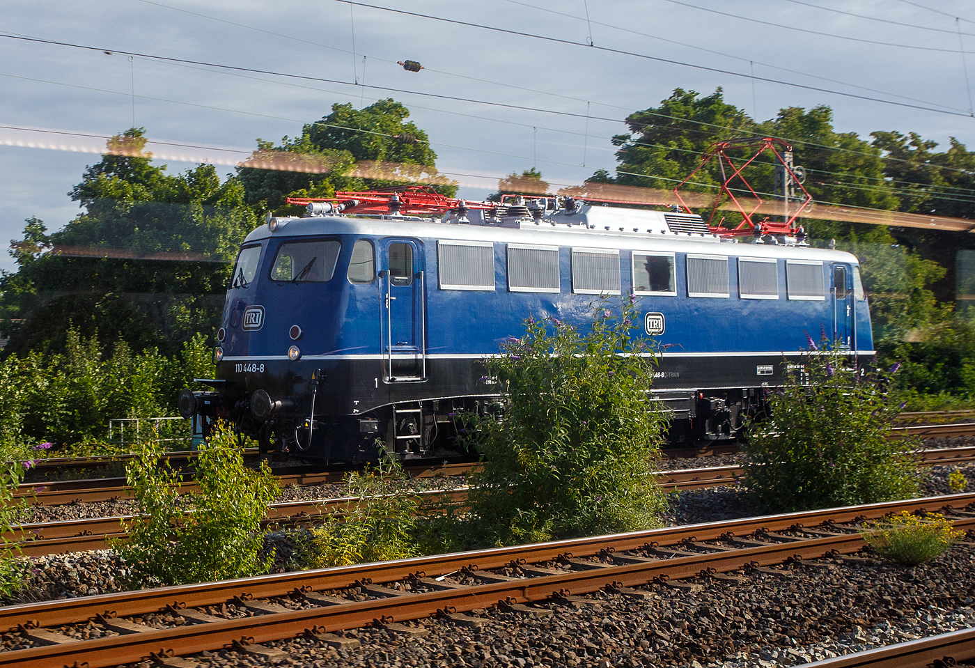 Auf unserer Fahrt mit einem IC nach Oberhausen am 24 Juni 2025 kommt uns kurz vor Oberhausen Hbf diese Bügelfalten E 10 entgegen, leider mit Spiegelungen da durch die Scheibe fotografiert. Es ist die 110 448-8 (91 80 6110 448-8 D-TRAIN) der TRI Train Rental GbR (Eckental-Eschenau), ex DB 115 448-3, ex DB 110 448-8, ex DB E 10 448-8.

Die E 10 (Bügelfalte ohne Verschleißpufferbohlen) wurde 1967 von der Krauss-Maffei AG in München-Allach unter der Fabriknummer 19223 gebaut, die Elektrik ist von den SSW - Siemens-Schuckert-Werke in Berlin. Sie wurde als E 10 448 an die Deutsche Bundesbahn geliefert. Mit der Einführung des EDV-Nummernsystems wurde sie zum 01.01.1968 zur DB 110 448-8, zum 17.12.2005 ging sie an die DB AutoZug GmbH und wurde so nun zur DB 1115 448-3 als solche kam sie 2015 zur DB Fernverkehr AG, am 17.01.2019 entgleiste die Lok beim Rangieren mit allen Achsen in Engelsdorf. So wurde sie 2020 ausgemustert und an die TRI - Train Rental International GmbH verkauft. Bis Ende 2024 wurde die Lok durch die Baltic Port Services GmbH in Mukran betriebsfähig aufgearbeitet und am 10.Dezember 2024 durch die TRI GmbH wieder in Betrieb genommen. Anlässlich der Untersuchung erhielt die Lok im Dezember 2024 eine Neulackierung in der Farbgebung in saphirblau mit silbernem Dach.

Bemerkungen/Besonderheiten: Konventionelle-Wendezug-Steuerung (36-poliges Kabel), Linienzugbeeinflussung (LZB); CIR-ELKE (Abk. Computer Integrated Railroading – Erhöhung der Leistungsfähigkeit im Kernnetz)
