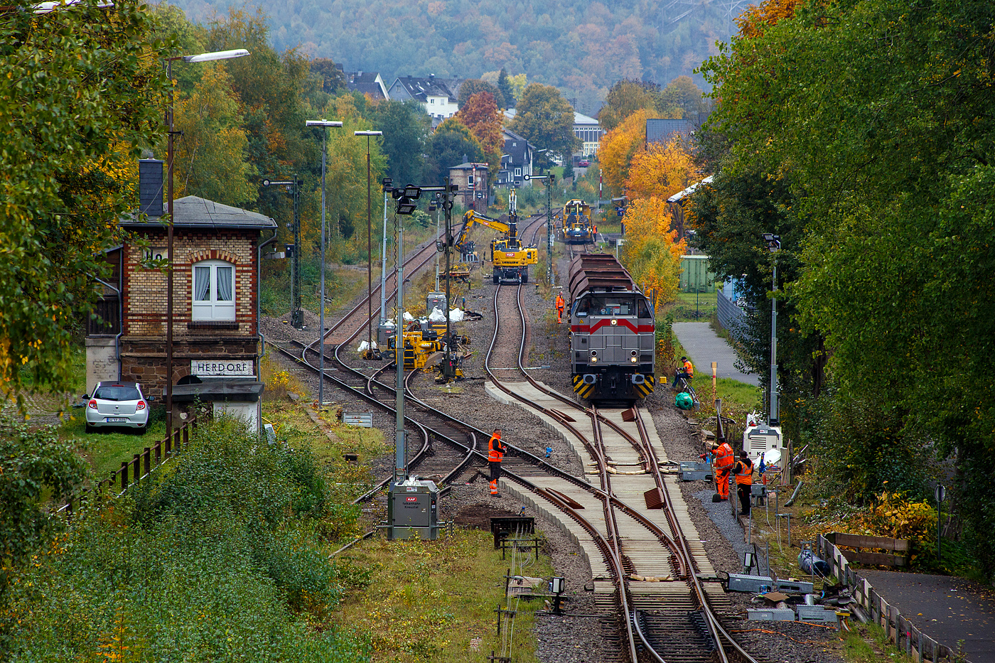 Blick auf den Bahnhof Herdorf und die Baustelle in Blickrichtung Betzdorf am 12 Oktober 2025. Die Weichen 25 und 26 sind beide eingebaut, müssen noch eingeschottert werden.

So steht die 277 809-0  Elmi“ (92 80 1277 809-0 D-KAF), eine Vossloh MaK G 1700 BB der KAF - Falkenhahn Bau AG (Kreuztal), steht mit einem Schotterzug bereit. Hinten beim Bahnhof haben die Schnellschotterplaniermaschine SSP 110 SW, Schweres Nebenfahrzeug Nr. 99 80 9425 068-0 D-DGU und die Universalstopfmaschine UNIMAT 09-475/4S, Schweres Nebenfahrzeug Nr. D-DGU 99 80 9424 001-2, den Bahnhof erreicht müssen aber nach das Einschottern abwarten. Beide Plasser & Theurer Maschinen gehören der Deutsche Gleisbau Union GmbH & Co. KG (DGU), Koblenz

Hier sieht man außerdem gut das Herdorf immer noch den Luxus von 2 Stellwerken besitzt. Vorne links das Weichenwärter Stellwerk Herdorf Ost (Ho) und hinten das Stellwerk Herdorf Fahrdienstleiter (Hf). Beide Stellwerke waren sogar hier bei der Baustelle ganztäglich besetzt.

Ich denke das bei den heutigen Verhältnissen im Bahnhof Herdorf, mit drei aktiven Gleisen und einer Anschlussstelle zum Rangierbahnhof der KSW Kreisbahn Siegen-Wittgenstein (Betriebsstätte FGE - Freien Grunder Eisenbahn) nicht mehr zeitgemäß ist. Es gibt mit Sicherheit heute Möglichkeiten die Aufgaben von einem oder dem anderen Stellwerk fernzusteuern. Früher mit bis zu 30 Gleisen und 3 Anschlussstellen musste es so sein, aber im heutigen Zustand