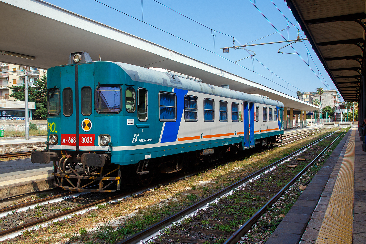 Der Dieseltriebwagen ALn 668 3032 (95 83 4668 332-8 I-Ti) der Trenitalia (100-prozentige Tochtergesellschaft der FS) steht am 19 Juli 2022, als Regionalzug als R 12946 nach Augusta, im Bahnhof Syrakus (Stazione di Siracusa). 

Der Dieseltriebwagen (Verbrennungstriebwagen) wurde 1980 von der Officine Meccaniche Calabresi O.ME.CA S.p.A. in Reggio Calabria gebaut und an die FS - Ferrovie dello Stato Italiane (Italienische Staatseisenbahnen) geliefert. Mit der Aufteilung in einzelne Betriebsgesellschaften der FS im Jahr 2000 kam der VT zur heutigen Tochtergesellschaft Trenitalia.

Die O.ME.CA. (deutsch Kalabrische Mechanische Werkstätten) war ein Hersteller von Schienenfahrzeugen mit Sitz in der Stadt Reggio Calabria, später gehörte sie zur AnsaldoBreda S.p.A., im Laufe der Zeit führte der Auftragsrückgang zu einer Verkleinerung der gesamten AnsaldoBreda-Gruppe und seit Februar 2015 gehört sie zur Hitachi Rail Italy S.p.A.. Die Stadt Reggio Calabria liegt an der Südspitze der italienischen Apenninhalbinsel (Stiefelspitze) und somit an der Ostseite der Straße von Messina gegenüber von Messina (Sizilien).

Geschichte der ALn 668:
Zu Beginn der 1950er Jahre war der Dieseltriebwagenbestand der italienischen Staatsbahnen für jene Dienste, für den er eigentlich ausgelegt war, praktisch unbrauchbar geworden. Zudem verursachte die große Zahl der verschiedenen Typen einige Schwierigkeiten beim Unterhalt und bei der Beschaffung von Ersatzteilen.

Zur Modernisierung des Reiseverkehrs auf dem sizilianischen Schmalspurnetz der FS wurden ab Januar 1950 Triebwagen des Typs RALn 60 eingesetzt. Bei der Konstruktion dieser Bauart konnten technische Erfahrungen gesammelt werden, die in der Folge auch für die regelspurigen (normalspurigen) Dieseltriebwagen von FIAT verwendet werden konnten.

Im Jahre 1954 wurde dann das Projekt eines standardisierten Dieseltriebwagens aufgenommen, welcher – gestützt auf die Erfahrungen mit den beiden bereits im Betrieb befindlichen Triebwagenserien – die Probleme im Dieselfahrzeugpark der FS lösen sollte. Der neue Triebwagen wurde auf 68 Sitzplätze ausgelegt, davon 8 erster und 60 zweiter Klasse. Gemäß dem Klassifikationssystem der FS wurde er als Leichter Dieseltriebwagen „Automotrice Leggera a Nafta 668“ (Leichter Dieseltriebwagen 668), kurz ALn 668 bezeichnet. Die Verdoppelung der Anfangsziffer „66“ wurde eingeführt, um Baureihen mit Vielfachsteuerung kenntlich zu machen.

Dank der breitgefächerten Verwendungsmöglichkeiten dieser Fahrzeuge und der mehr als 700 Exemplare, welche die italienischen Staatsbahnen in 30 Jahren anschafften, wurde diese Bauart vor allem ab den 1960er Jahren zum eigentlichen Rückgrat des italienischen Regionalverkehrs und erst in neuerer Zeit, durch die Minuetto, fast ganz auf Nebenbahnlinien verdrängt.

Projektierung und Anforderungen an das Fahrzeug:
Im Jahre 1954 bestellten die FS bei der Fiat Ferroviaria die ersten drei Prototypen der Serie: Sie erhielten die Nummern 1401 bis 1403.

Nach den gemischten Erfahrungen mit den großen einmotorigen Triebwagen ALn 990 und Aln 880 orientierte man sich beim Entwurf der neuen Fahrzeuge an die Erfolge mit den Vorkriegs-Triebwagen, den „Littorine“ (ALn 40, ALn 56, ALn 80 usw.), welche mit zwei Motoren ausgestattet waren. Es wurde auch der seit der Vorkriegszeit eingetretene technische Fortschritt mitberücksichtigt und die Technik gegenüber den Littorine wesentlich verbessert.

Die Entscheidung, die neuen Triebwagen mit zwei Motoren auszustatten, bot verschiedene Vorteile: Die beiden unabhängigen Traktionsgruppen mit weitverbreiteten Dieselmotoren erlaubten es, die Kosten für den Antrieb relativ tief zu halten, das Vorhandensein von zwei Treibachsen verbesserte die Adhäsion und die Ersatzteile waren günstiger und einfacher zu bevorraten. Alle mechanischen Teile sind einfach, leicht und wartungsarm ausgeführt. Zudem waren die Dieselmotoren aus der FIAT LKW-Massenproduktion was weiter die Kosten reduzierte.

Gegenüber den Vorkriegstriebwagen neu entwickelt wurde die Steuerung einschließlich der Kupplungsbetätigung, welche nunmehr elektrisch erfolgt. Damit konnte die von Straßenfahrzeugen bekannte Pedalbedienung aufgegeben werden. Ebenfalls eine Neuerung stellte die Verwendung von Lkw-Motoren für Triebwagen dar. Die wichtigste Neuerung betraf jedoch den Einbauort der Motoren. diese wurden nicht mehr im Kasten eingebaut, sondern befinden sich als „Unterflurmotor“ nunmehr unterhalb des Kastens. Damit konnten die Probleme der Instabilität bei hoher Geschwindigkeit und der Übertragung von Vibrationen des Zuges während der Fahrt auf heikle mechanische Teile, welche bei den Vorgängermodellen Quelle von Beschädigungen und häufigen Reparaturen war, gelöst werden.

Die Unterfluranordnung des Fahrmotors war auch das Merkmal der damals neuen Aln 668 Triebwagen, um maximalen Platz für Fahrgäste und Servicepersonal zu schaffen. Diese Technik setzte weltweit Maßstäbe im italienischen Eisenbahnbau, so dass der Einsatz des Horizontalzylindermotors Anfang der 1950er Jahre von den maßgeblichen internationalen Eisenbahnzeitschriften oft als „italienische Lösung“ bezeichnet wurde, die heute Standard ist (heute meist als Power-Packs).

Die Wagen erhielten Seitenpuffer und Regel-Schraubenkupplungen.

Von den Diesel-Triebwagen vom Typ FS ALn 668 wurden von 1951 bis 1981 insgesamt 787 Stück in 12 Serien bzw. drei Generationen gebaut. Mit der dritten Generation, der Serie 1200 und 3000 kehrten die FS wieder zu Mitteleinstiegen zurück, behielten jedoch die größere Wagenlänge der Serie 1900 bei. Durch einen vergrößerten Einstiegsbereich konnte die Ein- und Ausstiegszeit insgesamt verkürzt werden. Die Stirnseiten wurden verändert, das Dach ist an den Wagenenden nicht mehr heruntergezogen. Von dieser Dieseltriebwagen Serie vom Typ FS ALn 668.3000 (ALn 668 Serie 3000) wurden zwischen1980-1981 von FIAT Ferroviaria Savigliano und Officine Meccaniche Calabresi (O.ME.CA), insgesamt 40 Stück, gebaut.

FS Aln 668 – Serie 3000 – der 3.Generation:
Die FS ALn 668.3000-Triebwagen, auch bekannt als ALn 668 Baureihe 3000, gehören zur dritten Generation der ALn 668-Gruppe, die in chronologischer Reihenfolge aus den Baureihen 1900, 1000, 1200, 3000, 3100 und 3300 besteht.

Die Baureihe/Serie 3000 (FIAT Typ 7241 S) ist die aufgeladene Version der Baureihe 1200 (FIAT Typ 7241), die sich durch ihre Höchstgeschwindigkeit von 130 km/h, anstatt der 110 km/h, unterscheidet.

Bau der Serie 3000:
1978 wurden 120 Triebwagen des Typs ALn 668.3000 der FS bestellt. Die Serie blieb jedoch aufgrund der Entscheidung, die Mehrfachtraktionsmöglichkeit auf drei statt zwei Triebwagen zu erweitern, auf 40 Einheiten begrenzt. Diese Entscheidung führte 1980 zur Schaffung der neuen Serie 3100 mit den verbleibenden 80 Triebwagen.

Der Bau der vierzig Triebwagen des Typs ALn 668.3000 wurde dann zu gleichen Teilen zwischen FIAT Ferroviaria und Officine Meccaniche Calabresi (O.ME.CA), aufgeteilt. So wurden die ALn 668.3001-3020 von FIAT Ferroviaria in Savigliano (im Piemont) 1980 und die ALn 668.3021-3040 (wie dieser hier) von der O.ME.CA S.p.A. in Reggio Calabria 1980 und 1981 gebaut und geliefert.

Merkmale:
Die Triebwagen ALn 668.3000 verfügen über eine Kapazität von 68 Sitzplätzen, 8 Sitzplätze in der 1.Klasse und 60 (heute 57) in der 2.Klasse. Die Triebwagen und wurden von zwei Dieselmotoren mit einer Nennleistung von jeweils 147 kW angetrieben, die später auf 170 kW erhöht wurde. Diese ermöglichen eine Höchstgeschwindigkeit von 130 km/h und eine Reichweite von ca. 600 km. 

Die Triebwagen ALn 668.3000 können zusammen mit den Steuerwagen Ln 664.1400 gefahrenwerden und sind für den Mehrfachtraktionsbetrieb in Doppeltraktion geeignet.


Unterschiede zur 1200er-Serie: 
Der ALn 668.3000 unterscheidet sich von der 1200er-Serie nur durch den aufgeladenen IVECO 8217.32-Dieselmotor und die daraus resultierende Dimensionierung der Getriebekomponenten in Abhängigkeit von der höheren entwickelten Leistung, sowie höher Höchstgeschwindigkeit von 130 km/h.

Motor und Getriebe:
Der Motor der 3000er-Serie nutzt die aufgeladenen IVECO 8217.32-Motoren, die ab der 1900er-Serie verbaut wurden. Ihre gesteigerte Leistung ermöglicht eine Höchstgeschwindigkeit von 130 km/h ohne Achs-Getriebewechsel und mit der gleichen Achsübersetzung wie bei den 1900er- und 1000er-Serien.

Der aufgeladene Motor erforderte außerdem den Einsatz eines größeren Hydraulikgetriebes, einer Zweischeibenkupplung und die Verlegung der Schalldämpfer vom Dach in spezielle vertikale Auspufföffnungen an den Seiten des hinteren Vorraums, wie bei den Vorgängermodellen der 1900er- und 1000er-Serie.

TECHNISCHE DATEN der ALn 668 Serie 3000 (3. Generation):
Baujahre: 1980/81
Gebaute Anzahl: 40
Spurweite: 1.435 mm (Normalspur)
Achsfolge: (1A)(A1)
Länge über Puffer:  23.540 mm	
Drehzapfenabstand: 15.940 mm
Achsabstand im Drehgestell:  2.450 mm
Treib- und Laufraddurchmesser: 920 mm (neu)
Breite: 2.878 mm
Höhe:  3.698 mm
Eigengewicht: 37.000 kg
Motor (2 Stück): 6-Zylinder-Dieselmotor Typ IVECO 8217.32.033 
Hubraum: 13,8 Liter (Bohrung Ø137 mm x 156 mm Hub)
Nennleistung: ursprünglich 147 kW, später 170 kW bei 1.850 U/min (340 KW Ges.)
Leerlaufdrehzahl: 650 U/min
Ladeluftversorgung: Holset Turbolader (Cummins) mit Ladeluftkühler und Abgasrückführung
Hydrostatische: Kühlerlüftersteuerung
Durchmesser Drehmomentwandler (hydrodynamisch): 500 mm
Zweischeibenkupplung: Trockene Doppelscheibenkupplung
Übersetzungen FIAT 5-Gang-Schaltgetriebe (i=1: x): 5,37 im 1.Gang; 3,4 im 2.Gang; 2,01 im 3.Gang; 1,27 im 4.Gang und 1,0 im 5.Gang.
Maximale Geschwindigkeiten: 24 km/h im 1.Gang; 40 km/h im 2.Gang; 65 km/h im 3.Gang; 104 km/h im 4.Gang und 130 km/h im 5.Gang.
Installierte Leistung: 2 x 206 kW
Dauerleistung: 2 x 147 kW
Höchstgeschwindigkeit: 130 km/h
Leitungsübertragung: Jeweils vom Dieselmotor über hydrodynamischen Drehmomentwandler (hydraulisches Getriebe), Doppelscheibenkupplung, FIAT mechanisches 5-Gang-Schaltgetriebe und Achsengetriebe (i=1:2,39) auf die Achse und somit auf die Räder
Sitzplätze: 8 in der 1. Klasse und 57 in der 2. Klasse
Dieseltank (zweiteilig): 2 x 300 Liter = 600 Liter dm³ Tank
Stromgenerator: 2,4 kW 24 V Lichtmaschine
Luftkompressor Typ: AC.75ZB
Bremse: Frenu WU-P (42 t)

Die Nennleistung der Motoren war zunächst wie bei den Baureihen 1900 und 1000 auf 147 kW begrenzt (gedrosselt). Nach den positiven Ergebnissen der Tests mit den ALn 668.1000 und 3000 im Depot Cuneo sowie mit den ALn 668.3100 im Depot Benevento zwischen 1980 und 1981 beschlossen die Triebwagendepots jedoch, die Leistung auf 170 kW zu erhöhen.

Einsatz: 
Seit den neunnziger Jahre konzentrierten sich die Einsätze der ALn 668.3000er auf Sizilien im Depot von Palermo, von wo aus einige Einheiten im folgenden Jahrzehnt in die Depots nach Catania und Fabriano verlagert wurden.

Für den historischen Fuhrpark erhaltene Einheiten:
Im Jahr 2023 wurden die ALn 3010 und 3040 von der Fondazione FS Italiane für den Einsatz in historischen Zügen erworben und erhielten ihre ursprüngliche „graublau-beige Pergament“-Lackierung zurück, sie sind derzeit im Lokdepot Palermo untergebracht. Ansonsten sind sie seit Juli 2025 wohl nicht mehr im Einsatz.

Trotz ihres Alters waren die ALn 668 aufgrund ihrer niedrigen Betriebskosten sowie ihres hohen Komforts und ihrer Zuverlässigkeit von ihrer Bauzeit bis Juli 2025 im Einsatz. Sie wurden nun durch modernere und umweltfreundlichere Züge wie den dieselbetriebenen Minuetto ALn 501/502, die Mehrstoff-Elektrozüge Blues HTR 312/412, den Pesa ATRIBO ATR 220 und verschiedene Stadler-Straßenzüge wie den ATR 115 und den ATR 803 ersetzt.

Der ALn 668 galt als Standardtriebwagen mit Verbrennungsmotor der FS. Der ALn 663 ist völlig ähnlich und erhielt trotz gleicher Mechanik eine andere Klassifizierung (BR), die sich ausschließlich auf die neue Innenausstattung stützte, die die Anzahl der Sitzplätze von 68 auf 63 reduzierte.

Quellen: Trenitalia, Anschriften am VT, Wikipedia (Italien und D) 
Stand: September 2025
