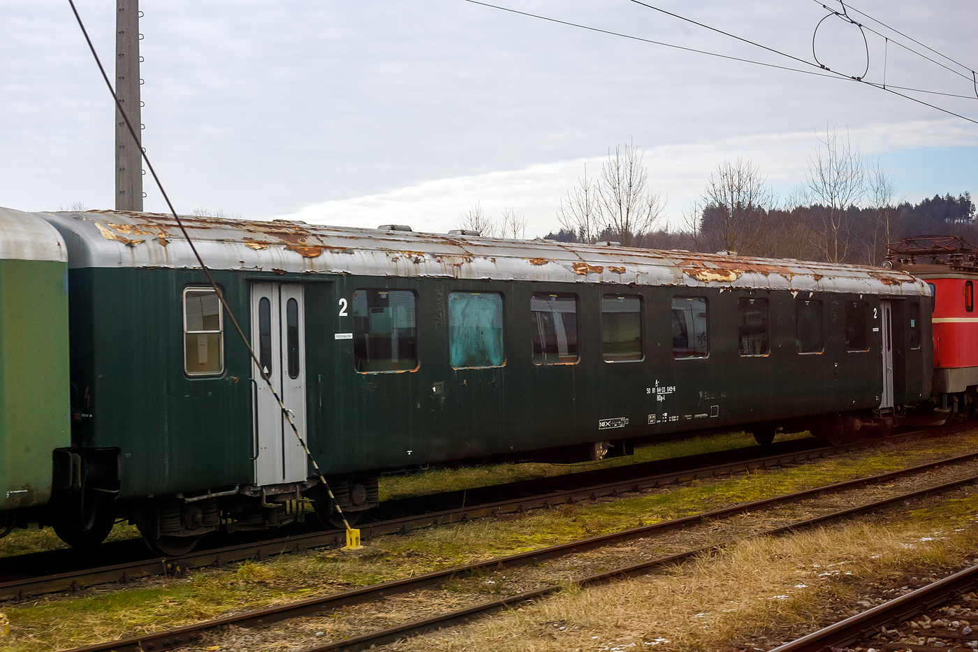 Der ex �BB Schlierenwagen der 2. Klasse Gro�raumwagen mit Mehrzweckabteil noch in Tannengr�n,  50 81 84-33 049-8 BDp-l der �GEG - �sterreichische Gesellschaft f�r Eisenbahngeschichte GmbH ist am 14 Januar 2025 beim Bahnhof Timelkam abgestellt. Bild leider nur aus einem Zug, h�tten wir gewusst was hier alles herumsteht, dann h�tten wir einen Stopp eingelegt.

Als Schlierenwagen wird eine Bauart von Reisezugwagen der �BB bezeichnet, die in den Jahren 1965 bis 1981 als Lizenzbau der Schweizer Einheitswagen I entstand. Der Name leitet sich von der Schweizer Stadt Schlieren ab, wo eine der beiden Lizenzgeberinnen ihren Sitz hatte. Die Schlierenwagen wurden bei den �BB bis Ende 2010 ausgemustert.

Es handelt sich um vierachsige Gro�raumwagen f�r den Inlandverkehr, bestellt im Jahr 1963 und gebaut von 1965 bis 1981 von SGP und den Jenbacher Werken (JW) in Lizenz von der Schweizerischen Industrie-Gesellschaft Neuhausen am Rheinfall (SIG) und der Schweizerischen Wagons- und Aufz�gefabrik Schlieren (SWS). Im Gegensatz zu den Schweizer Wagen besitzen die 1.-Klasse-Wagen nur 7 statt 8 bzw. die 2.-Klasse-Wagen nur 9 statt 10 Fenster mit Sitzgruppen.