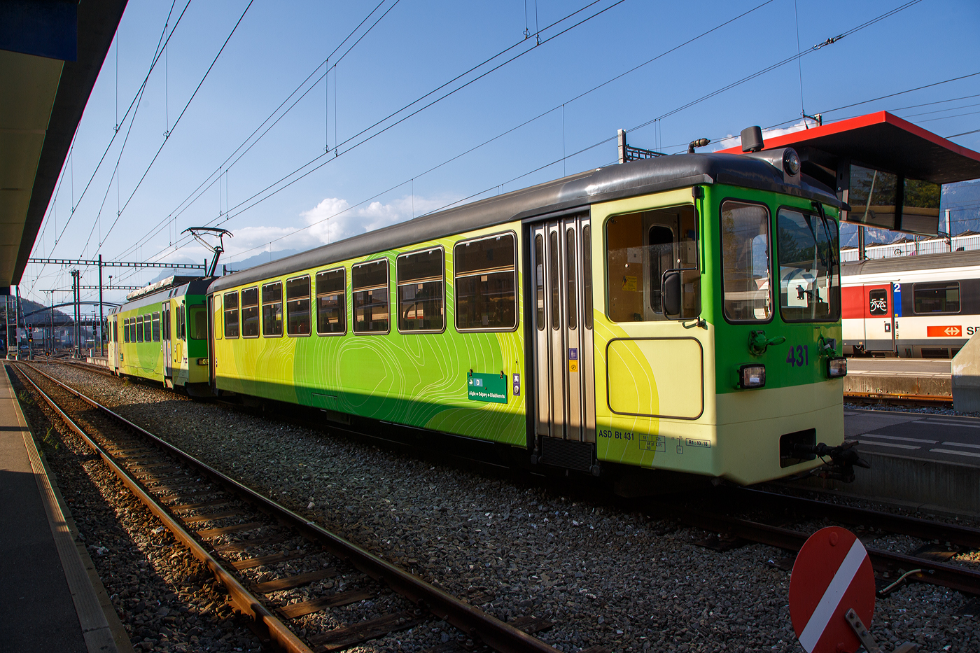Der Triebwagen tpc ASD BDe 4/4 403 „Ollon” mit dem Steuerwagen tpc ASD Bt 431 stehen am 10 September 2023, als ASD (Aigle–S�pey–Diablerets) Regionalzug R 24 nach Les Diablerets via Le S�pey, im  Bahnhof Aigle zur Abfahrt bereit. Die 23,3 Kilometer lange Strecke der ASD (Aigle–S�pey–Diablerets) ist eine reine Adh�sionsbahn und f�hrt von Aigle �ber Le S�pey nach Les Diablerets.

Der elektrische Personen-Triebwagen mit Gep�ckabteil wurde 1987 von Vevey ACMV (Ateliers de constructions m�caniques de Vevey) in Vevey gebaut, die elektrische

Ausr�stung ist von der BBC (Brown, Boveri & Cie.).  Er ist ein sogenannter ACMV Westschweizer Meterspurtriebwagen der zweiten Generation.

Die Westschweizer Meterspurtriebwagen sind elektrische Triebwagen, die 1985 bis 1992 von den Ateliers de constructions m�caniques de Vevey ACMV und 1996 von Vevey Technologies an sechs Westschweizer Privatbahnen f�r verschiedene Stromsysteme und zum Teil mit gemischtem Adh�sions- und Zahnradantrieb geliefert wurden. Die Fahrzeugfamilie ist modular aufgebaut und lie� sich bez�glich ihrer Gr��e und Ausstattung an die Erfordernisse der jeweiligen Bahngesellschaft anpassen. Die Triebwagen wurden elf Jahre produziert und w�hrend dieser Zeit der technischen Entwicklung angepasst.

Die Fahrzeuge sind in leichter Stahlbauart konstruiert und 2,65 Meter breit. Die Sch�rzen, der Unterteil des Kastens, wurden aus Aluminium-Stangpressprofilen hergestellt, die nach einer Kollision mit Kraftfahrzeugen ausgewechselt werden k�nnen. An den Stirnw�nden wurden bei weniger beanspruchten Elementen Formteile aus glasfaserverst�rktem Kunststoff verwendet. Die Wagenk�sten der Triebwagen sind f�r die Aufnahme der elektrischen Ausr�stung verst�rkt und mit Befestigungspunkten versehen. F�r die Laufdrehgestelle wurde eine Bauart �bernommen, die schon bei Steuerwagen der Chemin de fer Bi�re–Apples–Morges und Chemin de fer Yverdon–Ste-Croix verwendet wurde. Die Triebdrehgestelle verf�gen �ber einen Rolldrehkranz und zwei l�ngs angeordnete Fahrmotoren, die je eine Achse �ber Kardanwellen antreiben.

Neben einer Rekuperations- und einer Widerstandsbremse sind die Triebwagen mit einer selbstt�tigen Druckluftbremse ausger�stet. Weil deren Hauptleitung durch die Elektronik gesteuert wird, kann der Zug einh�ndig mit dem Fahrschalter bedient werden.

Die Aigle–S�pey–Diablerets-Bahn (ASD), seit 1999 Teil der Transports Publics du Chablais (TPC), konnten 1987 neues Rollmaterial beschaffen, so auch vier dieser Triebwagen BDe 4/4 (401 bis 404). Wegen ihrer engen Kurven dienten die kurzen NStCM-Triebwagen als Basisfahrzeug. Als Steuerwagen dienen der ASD Fahrzeuge der ehemaligen Birsigthalbahn, die sie von der Baselland Transport (BLT) erworben werden konnte. F�r den Einsatz auf der ASD wurde der F�hrerstand, bei den ex BTB-Steuerwagen neu aufgebaut.

Auch die AOMC (Aigle-Ollon-Monthey-Champ�ry-Bahn) hat drei solcher Triebwagen (BDeh 4/4 501 bis 503) beschafft, diese waren aber durch den Zahnradbetrieb und das andere Stromsystem der AOMC doch anders ausgef�hrt, u.a. auch in besonderer Leichtbauweise. Um die technischen Normalien ihrer Strecken zu vereinheitlichen, bauten die TPC im Jahr 2016 die Linie von Aigle nach Champ�ry um. Sie ersetzten das bisherige Zahnstangensystem Strub durch eine Abt-Zahnstange und erh�hten die Fahrleitungsspannung auf 1500 Volt. Damit verloren diese BDeh 4/4 501–503 und die zugeh�rigen Steuerwagen ihr Einsatzgebiet.

TECHNISCHE DATEN:
Baujahr: 1987 (4 St�ck)
Spurweite: 1.000 mm
Achsformel: Bo’Bo’
Wagenkastenmaterial: Stahl
L�nge �ber Puffer:18.800 mm
Breite: 2.650 mm
Gewicht: 32,4 t
H�chstgeschwindigkeit: 65 km/h (urspr�nglich 25 km/h)
Leistung: 820 kW
Stromsystem: 1500 Volt DC (Gleichstrom)
Elektrische Ausr�stung: Sch�tzensteuerung
Sitzpl�tze: 32
Kupplungstyp: BSi -Kompaktkupplung

Die BSI-Kompaktkupplung ist eine mechanische Vorrichtung zum Verbinden zweier Schienenfahrzeuge zu einem Zug. Sie �bertr�gt die Zug- und Druckkr�fte innerhalb eines Zugverbandes und l�sst sich automatisch kuppeln und entkuppeln. Sie findet �berwiegend bei Stra�enbahnwagen und Regionaltriebwagen der Eisenbahn Verwendung. Die Kupplung wurde vom ehemaligen Unternehmen Bergische Stahl-Industrie-Gesellschaft in Remscheid entwickelt, inzwischen Teil der Wabtec-Gruppe.

Der Steuerwagen tpc ASD Bt 431, ex BTB/BLT (Bt 26):
Die Geschichte:
Anl�sslich der Modernisierung des BTB-Rollmaterials und f�r die Bildung von die Betriebsabwicklung stark vereinfachenden Pendelz�gen wurden 1966 auch sieben Steuerwagen Bt 21 bis 27 beschafft. Diese standen jedoch lediglich sechs neuen Triebwagen ABe 4/4 11 bis 16 gegen�ber. Der zus�tzliche Steuerwagen wurde ab 1977 f�r einen Pendelzug mit den entsprechend angepassten Be 4/4 der Serie 8 bis 9 aus dem Jahre 1951 ben�tigt.

Die Steuerwagen entsprachen bez�glich des wagenbaulichen Teils so weit als m�glich den Motorwagen. Es waren zwei Fahrgastabteile (Raucher/Nichtraucher) vorhanden. Auf der f�hrerstandslosen Seite befanden sich die Plattformen mit den beidseitigen, pneumatisch bet�tigten Faltt�ren ganz am Wagenende, um im Wageninnern m�glichst viele Sitzpl�tze anordnen zu k�nnen. Als Drehgestelle kamen solche elastischer Bauart mit Flexicoil-Lagerung und Gummi-Zusatzfedern sowie Klotzbremse zum Einbau.

Der F�hrerstand entsprach jenem der Motorwagen (ABe 4/4). Er war von Anfang an so ausgelegt, dass auch die entsprechend angepassten Be 4/4 8 und 9 ferngesteuert werden konnten. Eine Stirnwandt�re erm�glichte das Mitf�hren von Zusatzwagen oder das Einreihen eines Steuerwagens in der Zugsmitte. 1972 wurde der Zugfunk nachger�stet.

Mit der Fusion der Basler Vorortsbahnen 1974 gingen alle sieben Steuerwagen, welche immer Seite Rodersdorf an die Z�ge gestellt wurden, an die neugegr�ndete Baselland Transport AG (BLT) �ber ,so wurde dieser zum BLT Bt 26.

Nach der Betriebsumstellung im Herbst 1984 verkaufte die BLT alle sieben Fahrzeuge in die Westschweiz. Die Bt 22, 23, 24 und 25 kamen als Bt 132, 133, 134 und 131 zur Schmalspurbahn Aigle–Ollon–Monthey–Champ�ry (AOMC).

Die Bt 21 sowie 26 und 27 fanden den Weg zur Aigle–S�pey–Diablerets-Bahn (ASD). Ab September 1985 wurden sie bei ACMV in Vevey f�r den Betrieb mit den f�r 1987 bestellten Triebwagen BDe 4/4 401 bis 404 hergerichtet. Die Anpassungen umfassten im Wesentlichen:
• Neuanstrich
• Verschlie�en der vorderen Stirnwandt�ren
• Einbau eines neuen F�hrertisches
• Anpassungen an den Steuerstromkreisen
• Anpassungen der elektrischen, pneumatischen und mechanischen Kupplungen (BSi)
• Einbau von Schienenbremsmagneten in beide Drehgestelle

Die Inbetriebsetzung erfolgte ab Juni 1987 als Bt 431 bis 433.

TECHNISCCHE DATEN bei Inbetriebsetzung (1966):
Spurweite: 1.000 mm
Typenbezeichnung: Bt
Anzahl Wagen: 7
L�nge �ber alles: 17.212 mm
Gr��te Breite: 2.500 mm
H�he �ber Dach: 3.450 mm
Drehzapfenabstand: 11.000 mm
Achsabstand im Drehgestell: 1.800 mm
Eigengewicht: 17.000 kg
Sitz-/Stehpl�tze: 64 / 66 (zus�tzlich 2 Klappsitze)
H�chstgeschwindigkeit: 65 km/h
Anschaffungskosten/Wg.: CHF 356.205,–

Abweichende heutige Daten:
Eigengewicht: 18 t
Sitz-/Stehpl�tze: 56/ 60
Max. Ladegewicht: 2,5 t (so m�sste er eigentlich BDt 431 hei�en)

Als vierter Steuerwagen stie� 2000 der jahrelang abgestellte und nie in Betrieb genommene Bt 131 der AOMC (ex BTB/BLT Bt 25) hinzu. Dieses Fahrzeug erfuhr dieselben Anpassungen wie die Bt 431 bis 433, wobei jedoch in Abweichung dazu die Stirnwandt�re belassen wurde.