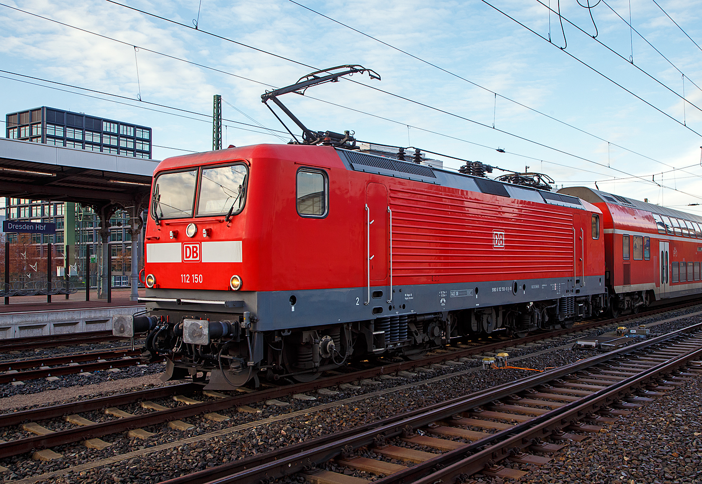 Die 112 150-8 (91 80 6112 150-8 D-DB) der DB Regio AG Nordost steht am 08.12.2022, mit einem Doppelstockzug im Hauptbahnhof Dresden.

Die Lok wurde 1992 von der AEG Schienenfahrzeuge Hennigsdorf GmbH unter der Fabriknummer 21487 gebaut und 1993 an die Deutsche Bundesbahn als DB 112 150-8 geliefert. Aus der Hennigsdorfer Lokschmiede LEW - VEB Lokomotivbau Elektrotechnische Werke „Hans Beimler“ Hennigsdorf wurde 1992 wieder AEG.

DB und DR bestellten jeweils 45 Lokomotiven der Baureihe 112.1. Die an die DR gelieferten Maschinen bekamen die Betriebsnummern 112 101-1 – 112 145-8, die an die DB gelieferten die 112 146-6 – 112 190-4. Nach dem Zusammenschluss der beiden deutschen Bahnen zum 01.01.1994 zur DB AG wurden aus den DR Maschinen DB.

Die 112.1 wurde unverhofft zu einem Symbol für die Deutsche Einheit, denn sie war die erste Lokomotivbaureihe, die von beiden Bahnverwaltungen gemeinsam beschafft wurde. Man kam überein, dass DR und DB jeweils 45 Loks der leicht verbesserten Loks der DR-Baureihe 112 bei der AEG bestellen würden (die AEG hatte zwischenzeitlich ihr 1946 enteignetes Werk in Hennigsdorf wieder übernommen). Dies erfolgte hauptsächlich aus arbeitsmarktpolitischen Gründen zur Stützung des Hennigsdorfer Werkes, denn die Bundesbahn hätte lieber eine 200 km/h schnelle und universell verwendbare Lok in aktueller Drehstromtechnik nach Art der Baureihe 120 beschafft.

Die äußerlich auffälligste Änderung ist die Zusammenlegung der großen Spitzen- bzw. Schlusslichter zu kleinen kombinierten Halogenlampen, so wie es bei der alten DB seit jeher gang und gäbe ist. 

Entwicklung:
Ab 1976 wurde von der Deutschen Reichsbahn in der DDR die Streckenelektrifizierung auf Grund der Ölverteuerung wieder forciert, nachdem man zehn Jahre zuvor noch ganz auf Diesellokomotiven gesetzt hatte. Der VEB Lokomotivbau Elektrotechnische Werke „Hans Beimler“ Hennigsdorf (LEW), einziger Hersteller von Elloks in der DDR, bekam den Auftrag, ausgehend von der bewährten schweren Güterzuglok der Baureihe 250 eine leichtere und technisch modernere vierachsige Variante für den Personenzug- und mittelschweren Güterzugdienst zu entwickeln. Diese sollte ohne betriebliche Einschränkungen die Lokomotiven der Baureihe 211 im Schnellzug- und die der Baureihe 242 im Güterzugdienst ersetzen können. Die mechanischen Komponenten wurden in Hennigsdorf entwickelt und hergestellt. Die Fahrmotoren kamen vom VEB Sachsenwerk Dresden.

Auf der Leipziger Frühjahrsmesse 1982 wurde der Prototyp 212 001 der Öffentlichkeit vorgestellt. Die Lokomotive machte vor allem durch ihr attraktives Äußeres (weiß mit roten Streifen) von sich reden und bekam von den Besuchern den Spitznamen Weiße Lady. Nach der Messe begann die DR mit der Erprobung der 140 km/h schnellen Lok, die konstruktiv für 160 km/h ausgelegt war. Nach der Probezerlegung im Ausbesserungswerk Dessau wurde die Getriebeübersetzung geändert und die Maschine am 14. Oktober 1983 in 243 001 umbezeichnet. Durch die geänderte Übersetzung konnte die Zugkraft deutlich erhöht werden, wobei die zulässige Geschwindigkeit auf 120 km/h reduziert wurde.

Serienfertigung DR-Baureihe 243, spätere DB-Baureihe 143
Da zu diesem Zeitpunkt (1984) im DR-Netz kaum mehr als 120 km/h gefahren werden durfte, wurden zunächst Mehrzwecklokomotiven der Reihe 243 beschafft. Von 1984 bis 1990 wurden 640 Loks gebaut.

Die DR-Baureihe 212, spätere DB-Baureihe 112.0
Da man bei der DR bis zuletzt davon ausgegangen war, dass der Ausbau der Transitstrecken nach West-Berlin und deren Elektrifizierung durch die Bundesrepublik finanziert würde, wurde erst 1991 mit dem Ausbau bestimmter Streckenabschnitte für mehr als 120 km/h begonnen. Allerdings standen bei der Deutschen Reichsbahn keine entsprechend schnellen Lokomotiven zur Verfügung. Es mussten neue, schnelle Lokomotiven beschafft werden. Man erinnerte sich daran, dass der Prototyp 212 001(später 243 001) ursprünglich für 160 km/h ausgelegt worden war. Nach vier Vorserienlokomotiven (212 002 bis 212 005) folgte eine Auslieferung von 35 Serienlokomotiven, bereits als 112 006 bis 040 bezeichnet. 

Von 1992 bis1994 wurden dann 90 verbesserte Loks (je 45 DR und DB) der Baureihe 112.1 von AEG gebaut. Ab Anfang 2001 standen diesie 90 Maschinen (der BR 112.1) aufgrund von Anrissen in der Aufhängung der Schlingerdämpfer für den hochwertigen Reisezugdienst nicht zur Verfügung. Die zulässige Höchstgeschwindigkeit wurde auf 140 km/h herabgesetzt und alle Schlingerdämpfer untersucht. Darüber hinaus wurde die Prüfung der Dämpfer im Rahmen der regelmäßigen Instandhaltung vorgeschrieben. Ende Januar 2001 standen die ersten Maschinen wieder mit einer zulässigen Geschwindigkeit von 160 km/h zur Verfügung.

Die 112.1 fand nach Überwindung einiger Kinderkrankheiten ihr Hauptbetätigungsfeld in der neu geschaffenen Zuggattung Interregio, aber auch den einen oder anderen InterCity bespannte sie. Durch den Wegfall der Gattung IR und den Ausbau des RE-Netzes mit 140-km/h- und 160-km/h-fähigem Wagenmaterial sind die Lokomotiven heute vorwiegend im Regionalverkehr anzutreffen. Die DB Fernverkehr gab deshalb zum 1. Januar 2004 alle ihre 112.1 an die DB Regio ab. 

DB-Baureihe 114.0
Alle Lokomotiven der Baureihe 112.0 wurden 2000 in die Baureihe 114 umgezeichnet, weil sie fortan zum Bestand von DB Regio gehörten, die Baureihe 112.1 verblieb ja noch beim Fernverkehr. Somit wollte man Verwechslungen zwischen den beiden Serien und damit auch zwischen Nah- und Fernverkehr vermeiden. Als die Fahrzeuge der Baureihe 112.1 auch in den Bestand von DB Regio eingereiht wurden, ist allerdings keine Rückumzeichnung der Baureihe 114 vorgenommen worden. 

TECHNISCHE DATEN der BR 112.1:
Hersteller: 	AEG
Baujahre: 1992 bis 1994 
Ausmusterung: seit 2019
Spurweite: 1.435 mm (Normalspur)
Achsformel: Bo’Bo’
Länge über Puffer: 16.640 mm
Drehzapfenabstand: 8.400 mm
Achsabstand im Drehgestell: 3.300 mm
Höhe: 3.980 mm
Breite: 3.120 mm
Dienstgewicht: 82,5 t
Höchstgeschwindigkeit: 160 km/h
Stundenleistung: 4.220 kW
Dauerleistung:  4.000 kW
Anfahrzugkraft:  226 kN
Stromsystem: 15 kV, 16,7 Hz~
Anzahl der Fahrmotoren: 4
Antrieb: LEW-Kegelringfeder
Bremse: KE-Druckluftbremse; elektrische Widerstandsbremse, 2.200 kW Dauerleistung (kurzzeitig bis 2.690 kW belastbar)
Zugbeeinflussung: LZB I 80
