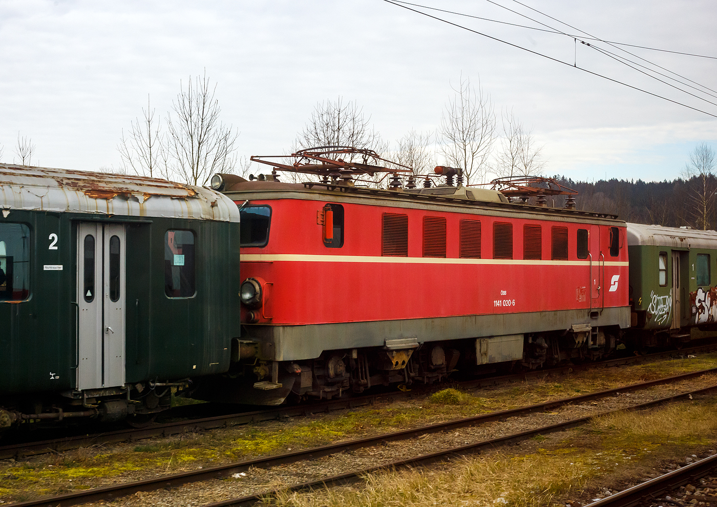Die 1141.20 (91 81 1141 020-6 A-�GEG) der �GEG - �sterreichische Gesellschaft f�r Eisenbahngeschichte GmbH, ex �BB 1141 020-6, ex �BB 1141.20, ist am 14 Januar 2025 beim Bahnhof Timelkam abgestellt. Bild leider nur aus einem Zug, h�tten wir gewusst was hier alles herumsteht, dann h�tten wir einen Stopp eingelegt.

Die vierachsige elektrische Universallokomotive 1957 von der Simmering-Graz-Pauker AG (SGP) in Graz, unter der Fabriknummer 56361, gebaut, bei dieser Lok ist die elektrische Ausr�stung von AEG-Union. Als �BB 1141.21 wurde sie an die �sterreichische Bundesbahnen geliefert. 1986 f�hrten auch die �BB EDV-gerechte Triebfahrzeugnummern ein. Aufgrund des zukunftssicheren Bezeichnungssystems waren die �nderungen �berschaubar. Die Ordnungsnummern wurden mit einer vorgesetzten Null dreistellig, der Punkt zwischen Stamm- und Ordnungsnummer entfiel und die Selbstkontrollziffern wurden erg�nzt, so wurde aus dieser die �BB 1141 021-4 als diese fuhr sie bis zu ihrer Ausmusterung im Jahr 2003 und wurde dann zum 01. Dezember 2003 an die �GEG verkauft..

Die Lokomotiven der Reihe 1141 der �sterreichischen Bundesbahnen (�BB) waren vierachsige elektrische Universallokomotiven die von 1955 bis 2003 im Einsatz waren. Die Maschinen wurden von Simmering-Graz-Pauker gebaut und stellen eine Weiterentwicklung der Reihe 1041 dar.

Die �sterreichischen Bundesbahnen ben�tigten nach dem Zweiten Weltkrieg neue Elektrolokomotiven f�r ihr umfangreiches Elektrifizierungsprogramm, womit die Dampftraktion abgel�st werden sollte. Da die Instandsetzung der wichtigsten �sterreichischen Bahnstrecken rasch voranschritt, wurden ebenfalls neue Lokomotiven ben�tigt. Der Bedarf wurde vorerst mit den Neubaulok-Reihen 1040 und 1041 gedeckt. Da sich die Loks der Reihe 1041 au�erordentlich gut bew�hrt hatten, entschieden sich die �BB f�r eine Weiterentwicklung, die als Reihe 1141 bezeichnet wurde. Als Mindestanforderungen legten die �BB eine H�chstgeschwindigkeit von 110 km/h, eine Dienstgewicht von maximal 80 t und eine Leistung von 3.000 kW fest; die Vorg�ngerreihe wies lediglich eine H�chstgeschwindigkeit von 90 km/h auf, weshalb sie bald vom Einsatz auf den Hauptstrecken verdr�ngt wurde. 

Zwischen 1955 und 1958 wurden 30 Maschinen an die �BB geliefert. SGP in Graz war f�r den mechanischen Teil zust�ndig, der elektrische Teil wurde von den Firmen AEG-Union, BBC, ELIN und SSW hergestellt. Alle Lokomotiven waren bei ihrer Ablieferung Tannengr�n lackiert, der Rahmen und das Laufwerk waren schwarz lackiert. Nach einigen Dienstjahren erhielten die 1141er ein blutoranges Farbkleid, einige erhielten sp�ter noch eine verkehrsrote Lackierung, auch wurden teilweise die F�hrerstandst�ren verblecht und nach hinten in den Maschinenraum versetzt.

Die Loks der Reihe 1141 wurden nach ihrer Ablieferung auf der Westbahn und auf der S�dbahn im Schnellzugverkehr eingesetzt. Sie wurden allerdings im Laufe der Zeit von der Reihe 1042 verdr�ngt. Zuletzt waren die 1141 in Attnang-Puchheim stationiert. Im Jahr 2003 schieden alle Lokomotiven dieser Reihe aus dem Bestand der �BB aus. Die letzten Vertreterinnen waren 1141 007, 020, 021, 022, 024 und 028. 

Mechanische Konstruktion:
Der Wagenkasten entsprach im Gro�en und Ganzen der Vorg�nger-Reihe 1041. Um die Lok so leicht wie m�glich zu konstruieren (die von den �BB gesetzte 80 t-Grenze durfte nicht �berschritten werden), wurden die Drehgestellrahmen in Holmenbauweise ausgef�hrt, zudem wiesen die Drehgestelle eine verbesserte Kurvenl�ufigkeit auf. Die Zugkraft�bertragung erfolgte auf gleiche Weise wie bei der Reihe 1041. Der aus Stahlblechen geschwei�te Kasten st�tzte sich auf Gleitbacken am Wiegebalkenende ab. Zwischen den beiden F�hrerst�nden lagen der Maschinenraum und ein Verbindungsgang, der Transformator befand sich in der Mitte der Lok. Bei den Lokomotiven der Baureihe 1141 wurde erstmals der heute bekannte und bew�hrte Siemens-Gummiringfederantrieb (ein Hohlwellenantrieb) eingebaut. Bevor die Reihe 1141 beschafft wurde, war dieser Antrieb nur probeweise in Bestandsloks verbaut. Die Reihe 1141 ist daher die erste in Serie gebaute Elektrolokreihe mit diesem Antrieb. Im Motorraum waren s�mtliche Hilfsaggregate und sonstige Ger�te untergebracht. Einige Lokomotiven erhielten im Zuge von Ausbesserungsarbeiten einen ver�nderten Kasten.

Elektrische Konstruktion:
Das Dach trug zwei Scherenstromabnehmer mit Doppelwippe (Bauart V), den Hauptschalter und s�mtliche Dachleitungen. Unter dem Hauptschalter befand sich der fremdbel�ftete und �lgek�hlte Transformator, der im Vergleich zur Reihe 1041 leicht ver�ndert wurde, die Steuerung wurde hingegen beibehalten. Die Loks verf�gten �ber vier Motoren, das �bersetzungsverh�ltnis des Getriebes betrug 23:88. Alle Lokomotiven verf�gten �ber eine automatisch wirkende Druckluftbremse, ein Nachbremsventil und eine direkt wirkende Zusatzbremse. Auf die Ausr�stung der Maschinen mit einer E-Bremse wie bei der Vorg�nger-Reihe musste aus Gewichtsgr�nden verzichtet werden.

TECHNISCHE DATEN:
Gebaute Anzahl: 30
Spurweite: 1.435 mm (Normalspur)
Achsformel: Bo’Bo’ (2 Drehgestelle mit je 2 Motoren)
L�nge �ber Puffer: 15. 260 mm
Drehzapfenabstand: 7.500 mm
Achsabstand im Drehgestell: 3.200 mm
Treibraddurchmesser: 1.350 mm (neu)
Dienstgewicht: 83,0 t
Stundenleistung bei 77 km/h: 2.480 kW
Dauerleistung: 2.160 kW
Stundenzugkraft: 116 kN
Anfahrzugkraft: 210 kN
H�chstgeschwindigkeit: 110 km/h
Anzahl der Motoren: 4 
Motorenart: 10-polige fremdbel�ftete Fahrmotoren (je 620 kW Stundenleistung)
Antrieb: Siemens-Gummiringfederantrieb
�bersetzung: 1: 3,83
Stromsystem: 15 kV/16,7 Hz