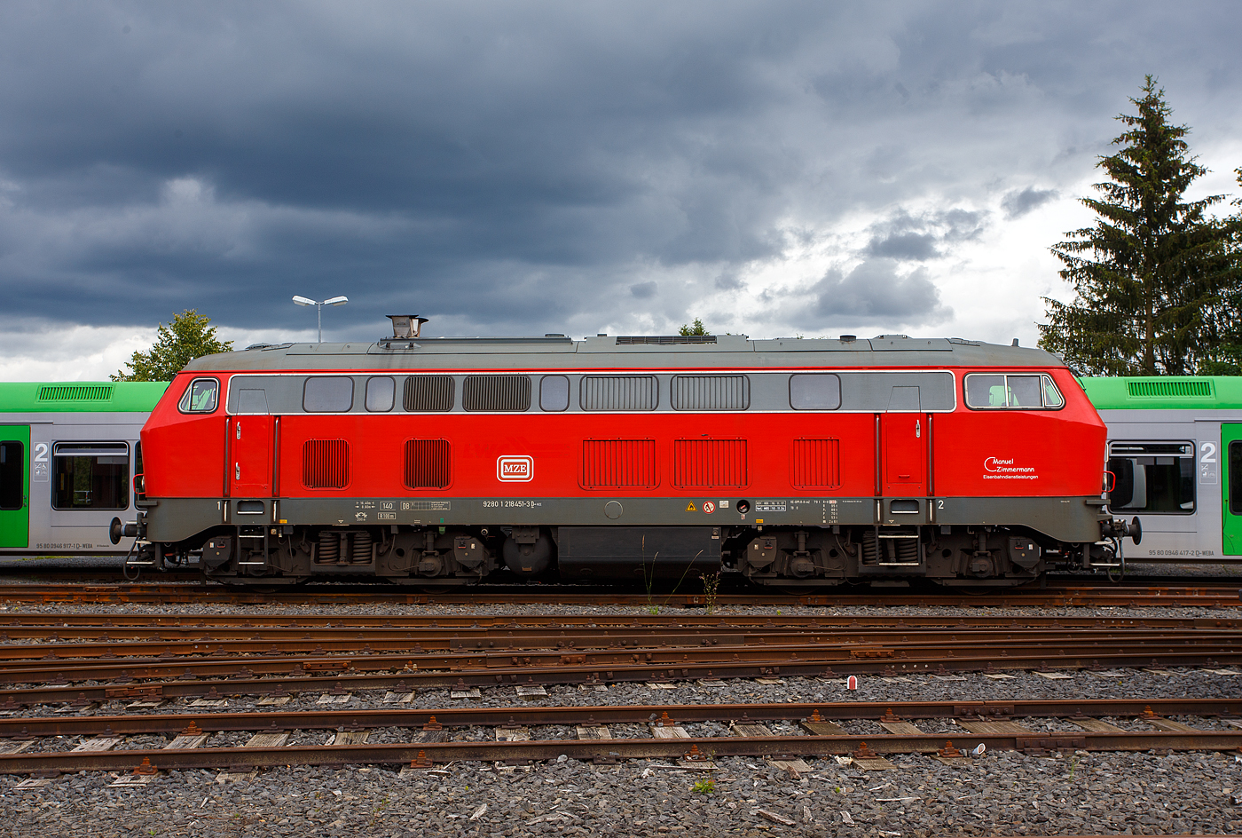 Die 218 451-3 (92 80 1218 451-3D-MZE) der MZE - Manuel Zimmermann Eisenbahndienstleistungen, Hellenhahn-Schellenberg (Ww), ist am 16 Juni 2024 auf dem Betriebshof der WEBA (Westerwaldbahn des Kreises Altenkirchen GmbH) auf der Bindweide bei Steinebach/Sieg abgestellt.

Die V 164 wurde 1978 bei Henschel in Kassel unter der Fabriknummer 32045 gebaut und als 218 451-3 an die DB geliefert. Bis April 2016 fuhr sie für die DB Regio AG – Nord als 92 80 1218 451-3 D-DB im Regionalbahn Einsatz in Niedersachsen und Bremen. Im Jahr 2016 ging sie dann an die LWC Lappwaldbahn Cargo GmbH in Braunschweig und war Ende 2021 an die Hering Bau GmbH & Co. KG in Burbach vermietet. Im Juni 2023 wurde sie dann an die MZE - Manuel Zimmermann Eisenbahndienstleistungen verkauft.

Die Baureihe 218 ist das zuletzt entwickelte Mitglied der V 160-Lokfamilie. Viele Gemeinsamkeiten der Baureihen V 160 bis V 169 (spätere 215 bis 219) wurden in ihr zusammengefasst. Bei der Baureihe 218 wurde von der Baureihe 217 die elektrische Zugheizung übernommen, von den Prototypen der Baureihe 215 übernahm man den 1.840-kW-Motor (2.500 PS), wobei ein Hilfsdieselmotor zum Betrieb des Heizgenerators überflüssig wurde. Später wurden noch stärkere Motoren mit 2.700 PS (KHD Lizenzbau Pielstick16 PA4 V 200) oder mit 2.800 PS (MTU 12 V 956 TB 11) verbaut. Den verbauten Motor kann man anhand der Anordnung der Abgashutzen auf dem Dach erkennen.

Im Jahr 1966 bestellte die Deutsche Bundesbahn zunächst zwölf Vorserien-Lokomotiven der Baureihe V164. Die ersten Lokomotiven wurden ab 1968 von der Firma Krupp ausgeliefert. Von der Deutschen Bundesbahn wurden sie aber als Baureihe 218 in Dienst gestellt. Die Serienbeschaffung (unter Beteiligung von Henschel, Krauss-Maffei und MaK in Kiel) erfolgte von 1971 bis 1979 mit 398 weiteren Maschinen. Hinzu kam 1975 nach einem Unfall die 215 112, die nach ihrer Instandsetzung zur 218 399 wurde. Die Auslieferung erfolgte in vier Bauserien (218 101-170, 171-298, 299-398, 400-499), bei denen es geringe Veränderungen gab.

Die 140 km/h schnellen und 2.500 bis 2.800 PS starken ''BB-Loks“  wurden im Reise - und Güterzugdienst eingesetzt. Die elektrische Zugheizung und die Wendezug- und Doppeltraktionssteuerung machen die Baureihe 218 zu einer universal verwendbaren Lok. Die Baureihe 218 bewährte sich im Betriebsdienst und galt noch bis ins Jahr 2000 als die wichtigste Streckendiesellok der Deutschen Bahn AG. Leider wurden immer mehr Leistungen im Personennahverkehr durch Triebwagen ersetzt. Dadurch und durch Betreiberwechsel waren immer mehr Lokomotiven der Baureihe 218 bei der Deutschen Bahn AG überflüssig geworden. Im Juli 2009 waren noch etwa 200 Exemplare im Einsatz. Eine echte Nachfolgerin für die Baureihe 218 ist noch nicht in Sicht.

TECHNISCHE DATEN:
Spurweite: 1.435 mm (Normalspur)
Achsformel:  B'B'
Spurweite:  1.435 mm
Länge: 16.400 mm
Breite des Lokkastens: 3.136 mm
Höhe des Lokkastens: 4.275 mm über Schienenoberkante
Drehzapfenabstand:  8.600 mm
Achsabstand im Drehgestell: 2.800 mm
Treibraddurchmesser: 1 000 mm (neu) / 920 mm (abgefahren)
Gewicht:  79 Tonnen
Achslast: 19,5 t
Motor: MTU wassergekühlter V12-Zylinder-Viertakt -Dieselmotor vom Typ 12 V 956 TB 11 (abgasoptimiert ) mit Direkteinspritzung, Abgasturboaufladung und Ladeluftkühlung
Motorleistung: 2.800 PS (2.060 kW) bei 1,500 U/min
Motor-Hubraum: 114,7 Liter
Anfahrzugkraft: 235kN 
Dauerzugkraft: 175kN
Höchstgeschwindigkeit:  140 km/h (im Langsamgang 100 km/h)
Motorleerlaufdrehzahl (ohne Zugheizung) : 630 - 650 U/min 
Motorleerlaufdrehzahl (mit eingeschl. Zugheizung): ca 1000 U/min 
Kleinste Dauergeschwindigkeit (im Schnellgang): ca. 28 km/h
Kleinste Dauergeschwindigkeit (im Langsamgang) ca. 18 km/h
Getriebe: MTU-Getriebe K 252 SUBB (mit 2 hydraulische Drehmomentwandler)
Leistungsübertragung: hydraulisch
Kleinster befahrbarer Gleisbogen: R = 100 m
Tankinhalt:  3.150 l
Sandvorrat: 400kg
Bremse: KE-GPP2R-H mZ
Bemerkung: Den verbauten Motortyp kann man auch anhand der Anordnung der Abgashutzen auf dem Dach erkennen.

Aufbau und Ausrüstung
Motor und Getriebe sind elastisch im Rahmen gelagert. Der Rahmen stützt sich über je vier Schraubenfedern pro Drehgestell und Seite auf die Drehgestelle ab. Ab 218 299 wurden dabei Flexicoil-Federn verwendet, die eine gleitstücklose Bewegung der Drehgestelle ermöglichen (Atlas-MaK-Drehgestelle). Zug- und Bremskräfte werden über tiefliegende Querträger mit Drehzapfen übertragen. Die Aufbauten sind in Schalenbauweise aus Blechen und Profilstahl gefertigt. Zwischen den beiden Endführerständen verlaufen zwei Seitengänge.

Der Antrieb erfolgt über vier sehr lange Gelenkwellen von einem zentral liegenden Strömungsgetriebe mit Wendegetriebe. Dieses Getriebe ist mit zwei Drehmomentwandlern ausgerüstet. Die Schaltung von Wandler I auf Wandler II erfolgt automatisch und ohne Zugkraftunterbrechung. Außerdem ist das Getriebe mit zwei Geschwindigkeitsstufen ausgerüstet. Die kleine Geschwindigkeitsstufe (Langsamgang) ermöglicht höhere Zugkräfte, allerdings eine niedrigere Endgeschwindigkeit von 100 km/h, die große Geschwindigkeitsstufe (Schnellgang) erlaubt die höhere Höchstgeschwindigkeit von 140 km/h, jedoch mit niedrigeren Zugkräften. Die Umschaltung von Langsam- auf Schnellgang erfolgt ausschließlich bei stehender Lokomotive. Zudem sind die Serienlokomotiven mit einer verschleißfreien hydraulischen Bremse ausgerüstet.

Im Laufe der Zeit wurden verschiedene Dieselmotoren als Antrieb eingesetzt: Die ersten Serien erhielten den MTU MA 12V 956 TB10 mit 2.500 PS. Um den bei diesen Motoren eintretenden Zugkrafteinbruch bei voller Heizleistung zu umgehen, wurden bei späteren Maschinen einige von KHD in Lizenz gebaute französische Pielstick 16 PA4 V200 mit rund 2,700 PS eingebaut. MTU zog nach und entwickelte aus dem TB10 den 2,800 PS leistenden stärkeren TB11, der in weit mehr als der Hälfte aller 218 zum Einbau kam. Durch schlechte Abgaswerte und fortschreitenden Verschleiß der Motoren sah sich der Betreiber in den letzten Jahren zu einem umfassenden Modernisierungsprogramm gezwungen. Zunächst wurden etliche TB11-Motoren mit einem Umrüstsatz abgasoptimiert. Weiterhin wurden mehrere diesem Stand entsprechende TB11-Motoren neubeschafft, die zudem über ein elektronisches Motormanagement verfügen. Parallel wurden auch gänzlich neue Motoren erprobt, was letzten Endes zur Beschaffung zahlreicher 16V 4000 R40/R41 von MTU führte. Außerdem wurden in drei Lokomotiven (218 329, 330 und 339) Motoren der Firma Caterpillar eingebaut. Durch den Zuwachs an Motoren wurde die Bestückung der Lokomotiven kräftig durchmischt. 

Ab 1981 wurden die Lokomotiven mit zusätzlichen Abgashutzen auf dem Dach ausgerüstet, um die Abgase an der Fahrleitung vorbeizuleiten und so deren Verschmutzung zu verringern, die durch Abgasklappen in den Hutzen erhöhte Austrittsgeschwindigkeit sollte zudem die Belästigung der Fahrgäste durch Abgase verringern. Die Anordnung der Hutzen ist ein Hinweis auf den in der Lokomotive eingebauten Motor: Lokomotiven mit Zwölfzylindermotor (TB10/TB11) haben entsprechend der einseitig am Motor angebrachten Abgasanlage nebeneinander angeordnete Hutzen, bei Sechzehnzylindermotoren (Pielstick, MTU 4000) mit Abgasanlagen an beiden Motorseiten sind die Hutzen versetzt angebracht.

