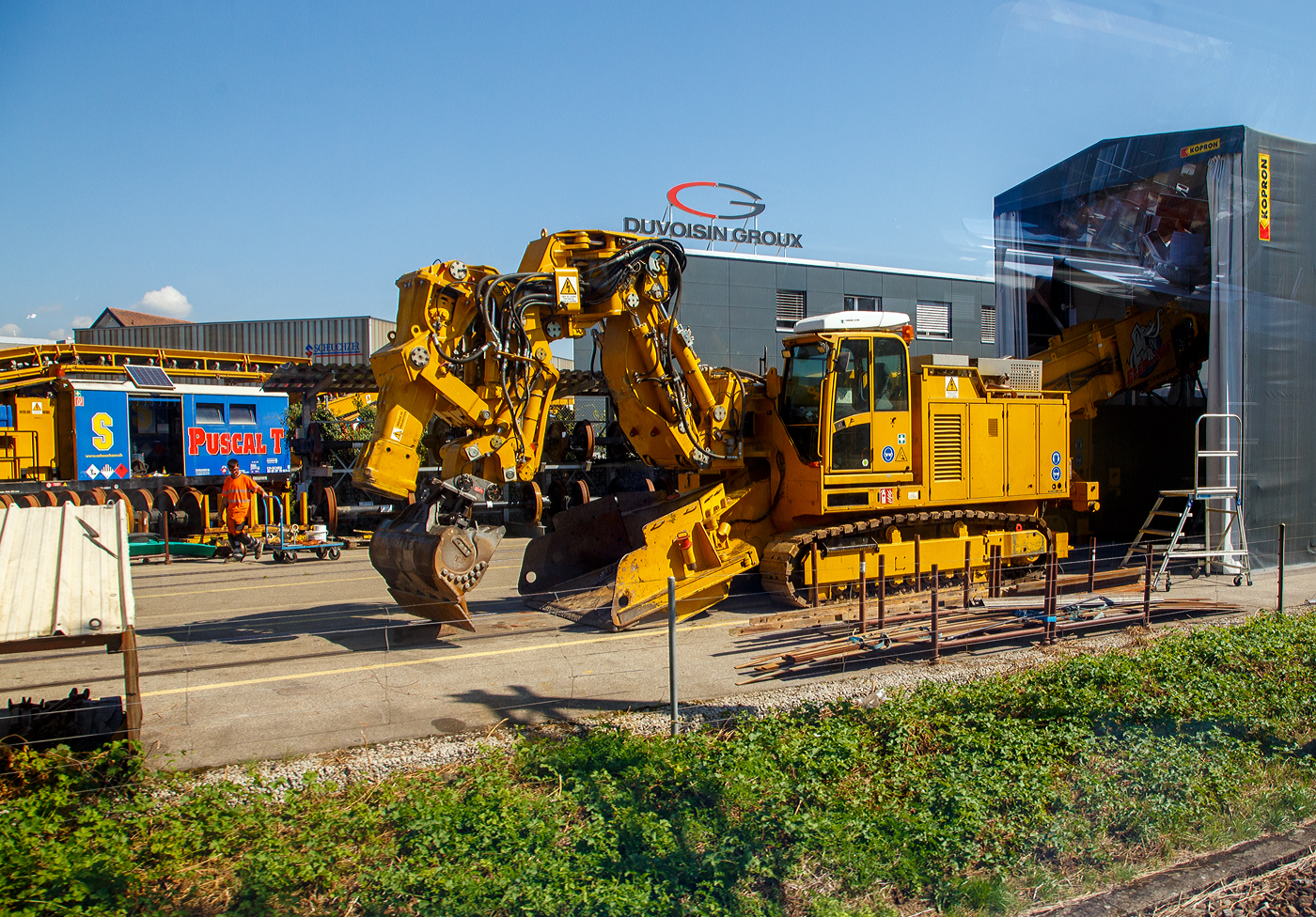 Ein Teil von dem PUSCAL T „Elefant“ der Scheuchzer SA am 07 September 2023 vor der Werkshalle der Scheuchzer SA in Bussigny (VD / Kanton Waadt). Bild aus einem Zug heraus.

Der Umbauzug Puscal T wird für die Unterbausanierung sowie den Wechsel der Schwellen und des Befestigungsmaterials eingesetzt. Die Maschine ist für den Felsabtrag, Tunnelabsenkungen oder das Abspitzen von fester Fahrbahn spezialisiert. Das Nachbargleis kann für den Zugsverkehr genutzt werden. Der Umbauzug ermöglicht den Abtransport des Aushubmaterials und die Zuführung der einzubauenden Materialien. Somit können die verschiedenen Untergrundschichten nach gewünschtem Profil (Geovlies, Kiessand, Splitt, Vorschotterung, Bitumen etc.) eingebaut, verteilt und verdichtet werden.

Hier im Vordergrund die Aushubmaschine „Elefant“ mit eine Spezialarm mit Baggerschaufel sowie integriertem 6 t Spitzhammer. Mit dieser ist eine Aushubtiefe bis zu 1.400 mm ab Schienenoberkante möglich. Ohne Spitzarbeiten ist ein Aushub von bis zu 100 m³/h möglich.
