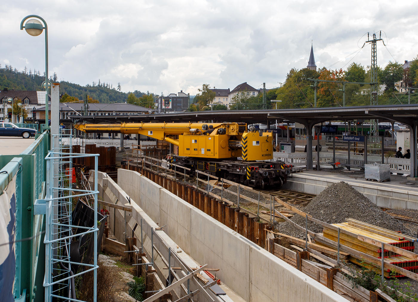 Nun im Einsatz der 125 t KIROW Gleisbauschienenkran KIROW Multi Tasker KRC 810 T (mit Schwerlastausr�stung), Schweres Nebenfahrzeug Nr. D-HGUI 99 80 9419 024-1, ex 97 82 53 513 19-3, der HERING Bahnbau GmbH (Burbach) ist am 29 September 2025 im Betzdorf/Sieg. 

Hier im Bereich unter dem P+R Parkhaus bis zum Fluss Sieg werden Horizontalbohrungen gemacht, vermutlich f�r erforderliche Entw�sserungs-Leitungen. So mussten Bohrrohre in die Baugrube gehoben werden und wie hier der Abraum aus der Baugrube gehoben werden. Eigentlich ist ein 125 t Kran damit unterfordert (�berdimensioniert), aber man muss bedenken es wurde eine weite Auskragung (Ausladung vor Puffer) und das freistehend (Nichtabgest�tzt) ben�tigt. 

Der hier mit der Ausladung vor Puffer von �ber 15 m k�nnte der Kran freistehend 30 t heben, aber darf dann nicht schwenken. Wenn geschenkt werden muss liegt diese dann nur bei 1,5 t. Entscheidend f�r die Ausladung des Auslegers ist aber auch die hintere Ausladung des Gegengewichts. 