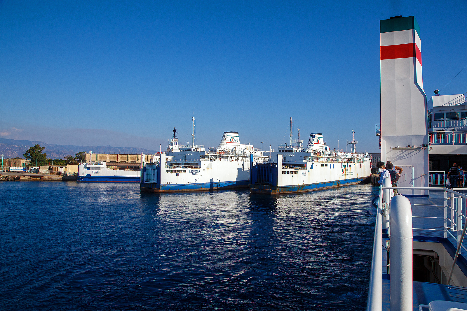 Schiffsbild ja, aber auch ein Bahnbild....
Zwei ungleiche Schwestern, die Eisenbahnfähren (nave traghetto) Scilla und Logudoro, der RFI Rete Ferroviaria Italiana (zur Gruppe der Ferrovie dello Stato Italiane) liegen am 20.07.2022 am Fährterminal von Messina Marittima. Wir sind mit dem mit unseren Trenitalia InterCity Notte ICN 1962, von Syrakus (Siracusa) nach Milano Centrale, auf der seit 2022 im Betrieb befindlichen RFI Neubau-Eisenbahnfähre Iginia II (Baujahr 2021) 

Vorne die Scilla (IMO 8219906), ihr Stapellauf war im September 1984. Dahinter die vier Jahre jüngere Logudoro (IMO 8506555) ihr Stapellauf war im Juni 1988. 

Das Hauptdeck jedes Schiffes hat 4 Gleise (ca. 400 m Gleis) für den Transport von Eisenbahnwaggons, während jeweils das Oberdeck vollständig für den Transport von Autos vorgesehen ist.

Die Logudoro unterschied sich ursprünglich von den Zwillingen Villa und Scilla, indem sie über einen Hotelservice verfügte, der für mehr als sieben Stunden Fahrt geeignet war. So hat sie auch mit zwei 8.400 kW starken Motoren anstelle der vier weniger leistungsstarken der Zwillinge. Zudem ist sie mit einen ausgeklügelten Stabilisierungssystem ausgestattet und so auch für offene See geeignet.