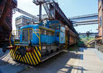 Die Dieselhydraulische Normalspur-Lokomotive Nr. 34, eine Henschel DHG 240 B  Esslinger , der Völklinger Hütte (ehem. Röchling'schen Eisen- und Stahlwerke GmbH in Völklingen/Saar, ab 1982 ARBED-Saarstahl GmbH, ab 1986 Saarstahl Völklingen GmbH) hier am 01 Juli 2025 abgestellt als Denkmal, mit zwei selbstentladende Schüttgutwagen der Gattung Fad oder Falns am Eingang der Rohstoffentladebunker in Richtung Hüttenbahnhof. Die Darstellung steht für den Rohstofftransport der Hütte zur Herstellung des Sintergutes. Die Lok war auch auf Bundesbahn Gleisen zugelassen. Heutiger Eigentümer der Lok und Wagen ist die Initiative Völklinger Hütte e.V..

Die zweiachsige dieselhydraulische Lok mit Antrieb über Gelenkwellen wurde 1969 von Henschel in Kassel unter der Fabriknummer 31210 gebaut und an die Röchling'sche Eisen- und Stahlwerke GmbH in Völklingen als Lok 34 geliefert. Weiterer Lebenslauf/Umfirmierungen: 
01.01.1971 > Stahlwerke Röchling-Burbach GmbH, Völklingen  34  
Juni 1982 	 > ARBED-Saarstahl GmbH, Völklingen  34  
Juni 1986 	 > Saarstahl Völklingen GmbH, Völklingen  34  
10.05.1989 > Saarstahl AG, Völklingen  34  
Später an MGS - Metallurgische Gesellschaft Saar mbH, Völklingen  34  
März 1999 	an Weltkulturerbe Völklinger Hütte - Europäisches Zentrum für Kunst und Industriekultur GmbH, Völklingen und Aufstellung als Denkmal. 

Die Lokomotive war bis zu einem Motorschaden 1997 in verschiedenen Bereichen eingesetzt: Transport von Duplexeisen zwischen der Mischerhalle und dem Siemens-Martin-Werk, im Bereich des Siemens-Martin-Werks, der Elektroöfen und des Hammerwerks sowie nach Einbau einer Funkfernsteuerung im Hafenbereich der Saarstahl AG.

Mit den Fahrzeugen der Typenreihe DHG 160 B / DHG 200 B / DHG 240 B und DHG 350 B bot Henschel Anfang der 1960er Jahre Gelenkwellenmaschinen für den unteren Leistungsbereich an, deren konstruktiver Ursprung nicht in Kassel, sondern in Esslingen der lag. Im Jahr 1961 übernahm Henschel das Diesellok-Typenprogramm der Maschinenfabrik Esslingen. Es wurde eine bei der Maschinenfabrik Esslingen als Typ Bern in Auftrag gegebene Lokomotive als DHG 325 in Kassel fertig gebaut und an den Auftraggeber, die Duisburger Kupferhütte, ausgeliefert. Der Typ Bern stellte auch die Basis für das oben genannte Typenprogramm dar. Dieses unterschied sich zwar optisch deutlich von seinem Esslinger Ursprung, übernahm aber die wesentlichen konstruktiven Merkmale der Fahrzeuge aus Esslinger Produktion. Gleichzeitig gab es keinen technischen und optischen Bezug zu den bisher im eigenen Henschel-Typenprogramm angebotenen Fahrzeugen.

Bei den neuen Gelekwellenfahrzeugen gab es leistungsmäßig deutliche Überschneidungen mit dem kleinsten Typ der 3. Generation, der stangengetriebenen DH 240 B. Diese wurde dann in der 4. Generation durch die DHG 240 B ersetzt.

Die Henschel DHG 240 B wurde zwischen 1963 und 1971 in 34 Exemplaren gebaut. Die Loks gingen vornehmlich an Chemiebetriebe und Raffinerien in Deutschland und der Schweiz. Von diesen wurden elf Loks explosionsgeschützt ausgerüstet. Einzelne Loks wurden ebenfalls mit Funkfernsteuerung ausgerüstet. Mit weitem Abstand am stückzahlenstärksten ist die DHG 240 B, die im Gegensatz zu ihren schwächeren  Schwestern  nicht mit einem DIWABUS-Getriebe von Henschel, sondern mit einem Voith-Hydraulikgetriebe ausgestattet ist. Zahlreiche Fahrzeuge stehen auch heute noch im täglichen Einsatz.

TECHNISCHE DATEN:
Baujahre: 1963 bis 1971
Gebaute Stückzahl: 34
Spurweite:	1.435 mm (Normalspur)
Achsfolge: B
Länge über Puffer: 7.160 mm
Achsabstand: 2.800 mm
größte Breite: 3.000 mm
größte Höhe über Schienenoberkante: 3.600 mm
Raddurchmesser: 850 mm (neu)
kleinster befahrbarer Gleisbogen: 35 m
Eigengewicht: 28 t
Max. Achslast: 16 t
Dieselmotor: wassergekühlter 6 Zylinder / 4-Takt-Diesel-Reihenmotor mit Turboaufladung, ohne Ladeluftkühlung vom Typ Henschel 6R 1416 A, Hubraum 13,3 l.
Leistung: 177 kW (240 PS)
Drehzahl: 1.600 U/min
Höchstgeschwindigkeit: 30 / 60 km/h
Leistungsübertragung: hydraulisch (Voit-Getriebe)
Kraftstoffvorrat: 400 l
